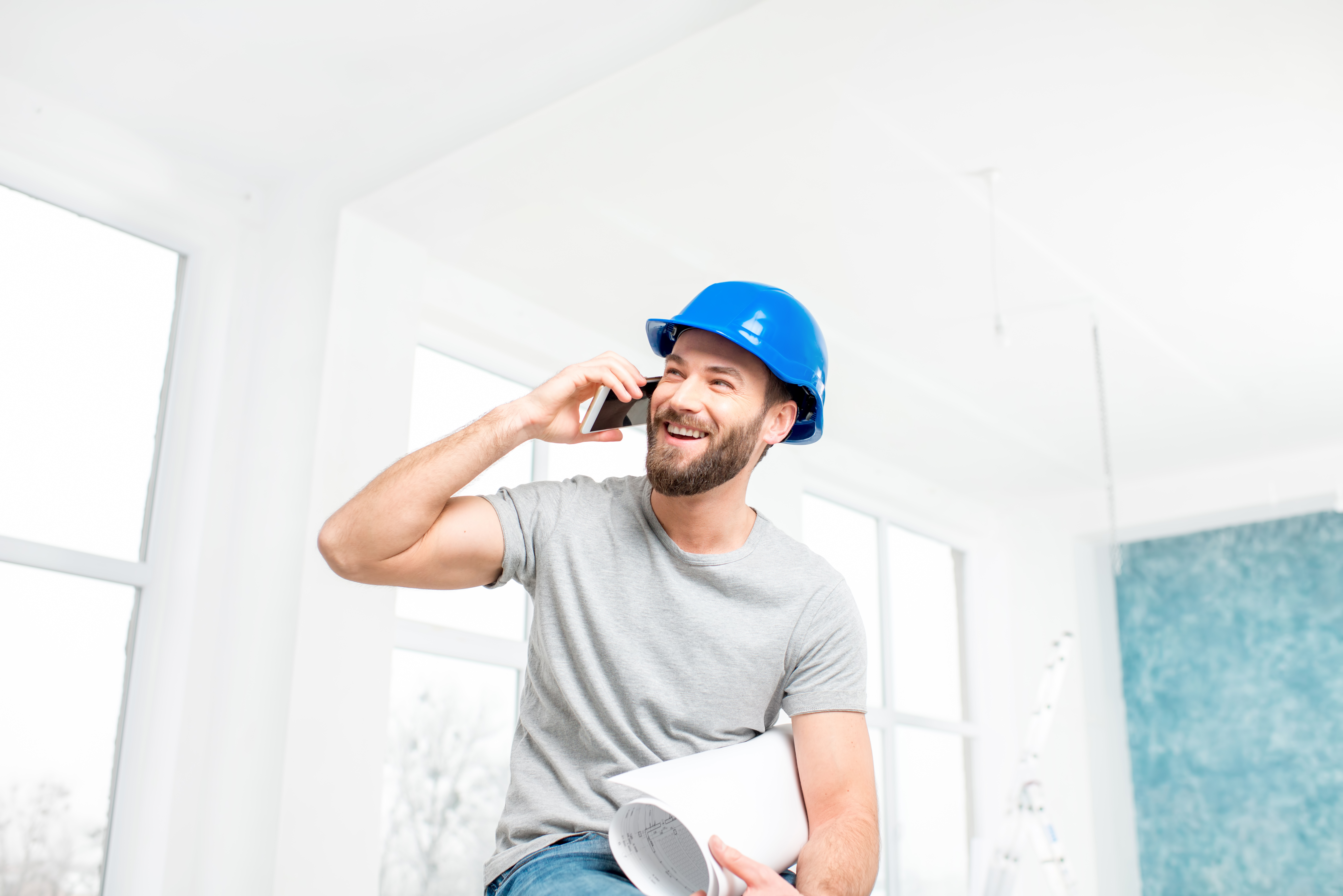 Handsome smiling repairman or builder in helmet talking with phone with drawings in the white interior